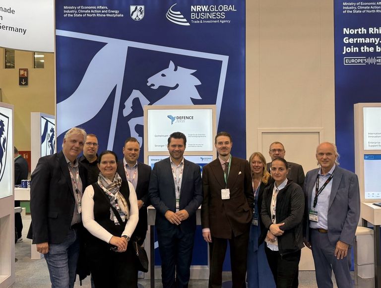 Gruppe von acht Personen bei einer Messe oder Konferenz vor Infostand.